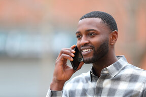 A smiling man with short hair is talking on his smartphone. He is wearing a checked shirt and appears engaged in the conversation. The background is blurred, focusing on his expression.