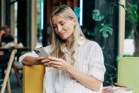 A woman with long blonde hair is sitting comfortably on a colorful chair, holding and looking at her smartphone. She wears a white shirt with stripes and appears engaged in her phone while in a casual café setting.