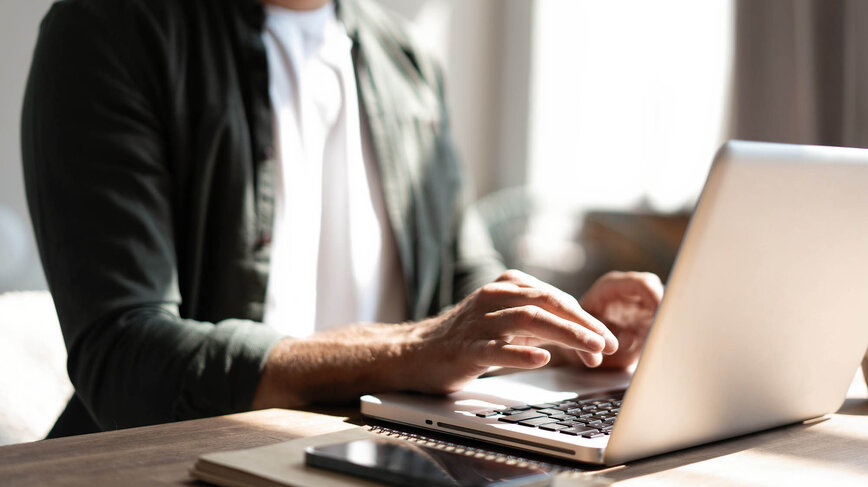 A person is using a laptop while seated at a table. Their fingers are positioned on the keyboard, and a smartphone is resting nearby. Soft natural light illuminates the scene, suggesting a peaceful indoor workspace.